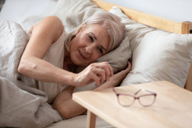 Mature woman taking glasses from bedside table in the morning Mature woman with eyesight problem taking glasses from bedside table in the morning after awakening, sleepy older female lying in comfortable bed at home, finding spectacles, bad vision concept squint eyes stock pictures, royalty-free photos & images