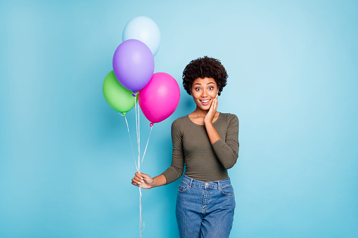 Portrait of amazed funky afro american girl hold colorful baloons receieved for her anniversary present from friends scream wow omg wear, good look outfit denim jeans isolated blue color background