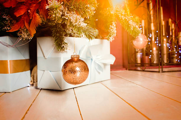 Xmas background mockup with copyspace. Boxes of gifs under cristmas fir tree on wooden floor near fireplace. Ornaments, garland and golden ball with beautiful orange backlight. stock photo