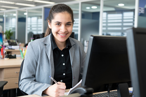 Young businesswoman at computer, with notepad and pen, looking towards camera with cheerful expression