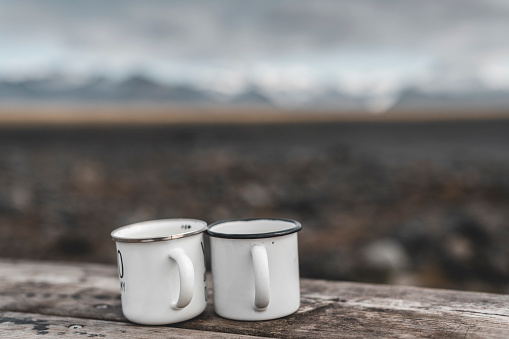 Two white enameled mugs on a wooden table. In the background the Icelandic landscape.