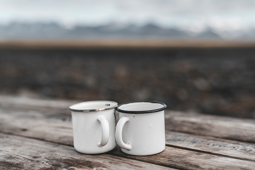 Two white enameled mugs on a wooden table. In the background the Icelandic landscape.