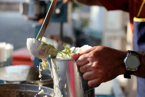 A closer look on senior man pouring coconut milk 'cendol' (green rice flour jelly droplets) into a mug of shaved ice at his business stall.