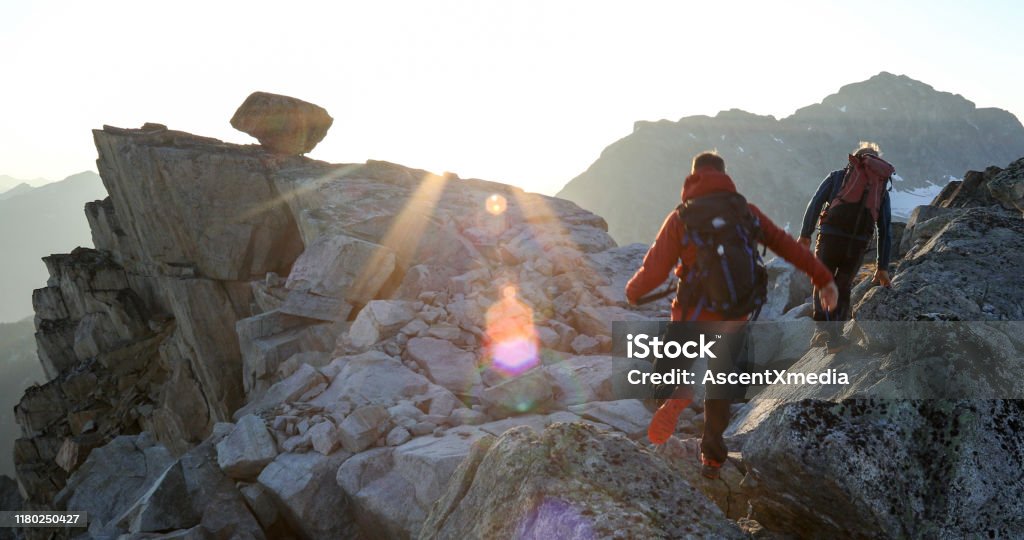 Two mountaineers traverse summit ridge at sunrise Snowcapped mountain ranges behind Hiking Stock Photo