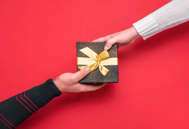 Giving and receiving festive gift box. Christmas gifting time concept. Man and woman hands on a black gift box on red background. Xmas gift exchange.