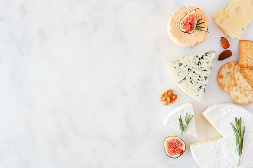 Border of a selection of cheeses, figs, nuts and crackers. Top view on a white marble background with copy space.