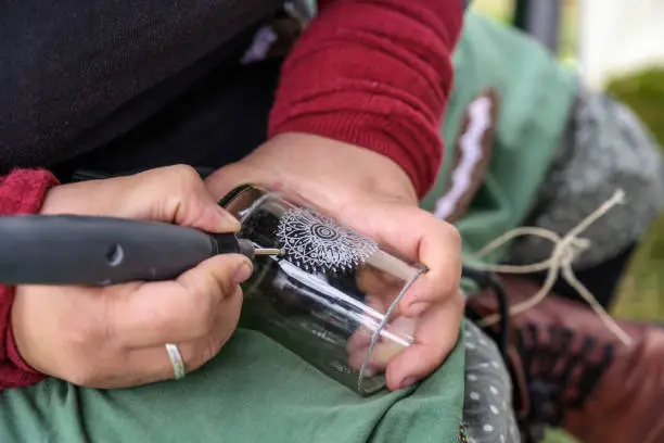 hands of a woman engraving a decorative pattern into a glass vase from a recycled bottle at a craft market, selected focus