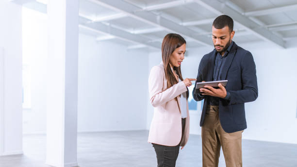 Asian businesswoman talking to interior designer in an empty office Asian businesswoman asking an African-American interior designer holding a digital tablet question on furnishing the empty office they are standing in while he is showing her options on a digital tablet. asian property stock pictures, royalty-free photos & images