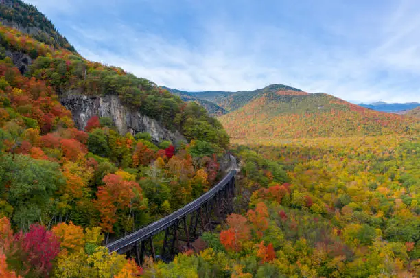 Aerial drone photo of during autumn day of the beautiful red, orange and yellow leaf foliage. Taken in the White Mountains, New Hampshire with train track trestle curving around mountainside.