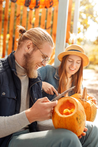 ハロウィーンプレパラトンコンセプト。ジャック・オー・ランタンを作る若いカップルは、楽しい側面のビューをクローズアップカボチャを彫ります - photography close up side view halloween ストックフォトと画像