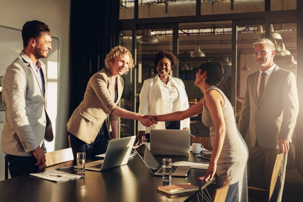 Two businesswomen shaking hands together after a successful boardroom meeting Two businesswomen shaking hands together over a table during a meeting with colleagues in a boardroom in the late afternoon merger stock pictures, royalty-free photos & images