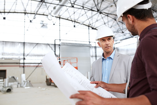 Two building engineers wearing hardhats and discussing blueprints while working inside of a large warehouse during an inspection