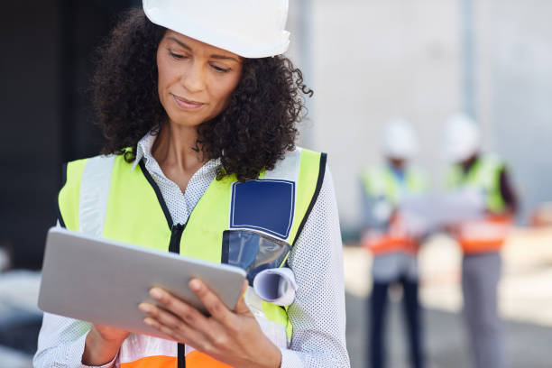 Female building engineer using a tablet on her work site Young female construction engineer wearing a safety vest standing outdoors on a worksite using a digital tablet with workers in the background occupational safety and health stock pictures, royalty-free photos & images