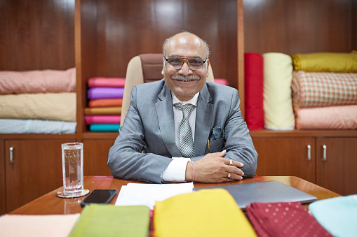 Successful Indian textile manufacturing CEO in mid 50s sitting at head of board room conference table surrounded by colorful textile samples.