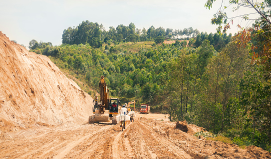 Nyungwe, Rwanda- May 2017: Locals riding their bicycles, while Heavy machines prepare the foundation of a new road construction in Rwanda, on a dirt road connecting Lake Kivu and Nyungwe.