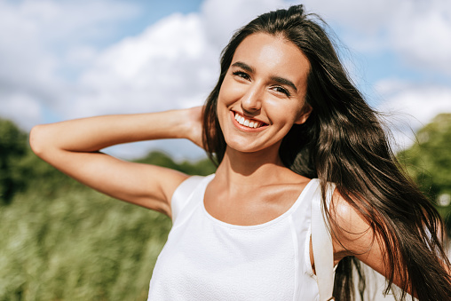 Happy young brunette woman smiling broadly with a windy blowing long hair in the park, posing on nature background.