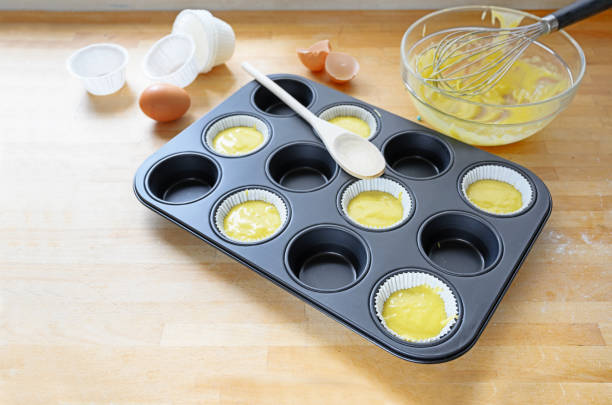 istock muffin tin, partly filled with paper cups and dough for cupcake baking on a wooden kitchen table, copy space 1180363349