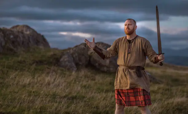 A lone redhead individual Scottish viking sword wielding warrior man wearing a kilt on highland moors