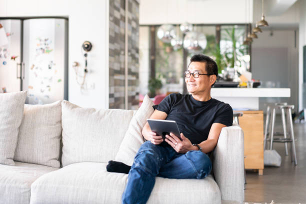istock Mature Chinese man sitting on sofa with a digial tablet in hands 1180378474