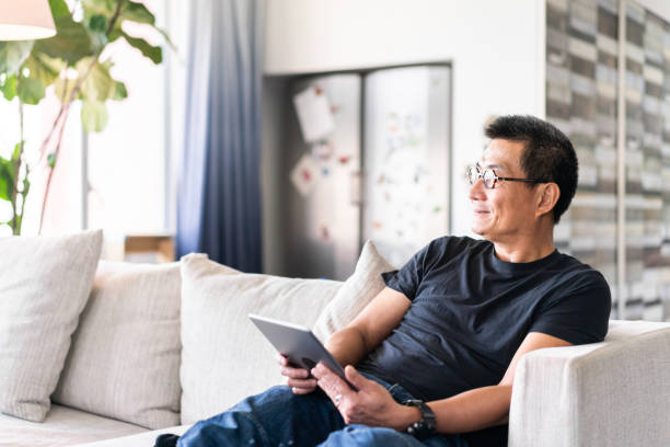 istock Mature Chinese man sitting on sofa with a digial tablet in hands 1180378633