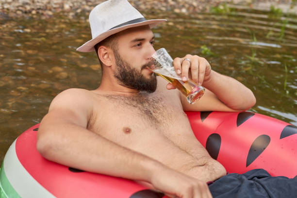 istock Young man with a beard and a hat sitting in a rubber boat and drinking beer from a transparent glass 1180379345