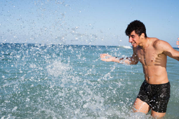 Young Caucasian woman goes at the sandy seaside in sunny day. Woman in shorts and striped t-shirt kicking sea water at the beach.Freedom and happy holidays vacations concept.Summer time, water splash.