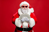 Portrait of cool stylish elderly santa claus hipster millionaire have cash, want buy presents spend on winter season sales discount wear cap hat eyewear eyeglasses isolated over red background