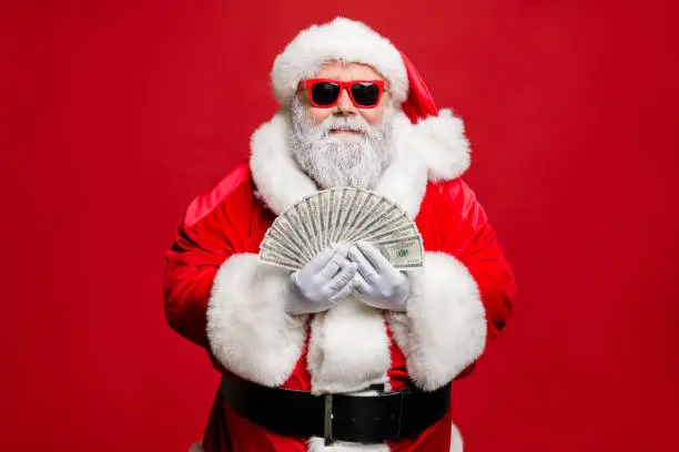 Photo of Portrait of cool stylish elderly santa claus hipster millionaire have cash, want buy presents spend on winter season sales discount wear cap hat eyewear eyeglasses isolated over red background