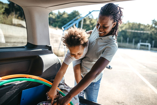 African woman packing stuffs in car trunk with little mischievous daughter