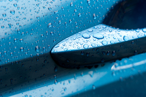 The door handle of a blue metallic colored car is wet after rain with droplets visible all across its surface.
