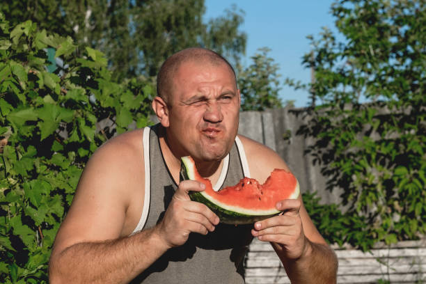 brutaler mann genießt süße rote wassermelone. 35-jährige männliche modell sitzt auf naturgrünen hintergrund und essen frisches obst. glücklich essen deich. - men 35 40 years male 30s stock-fotos und bilder