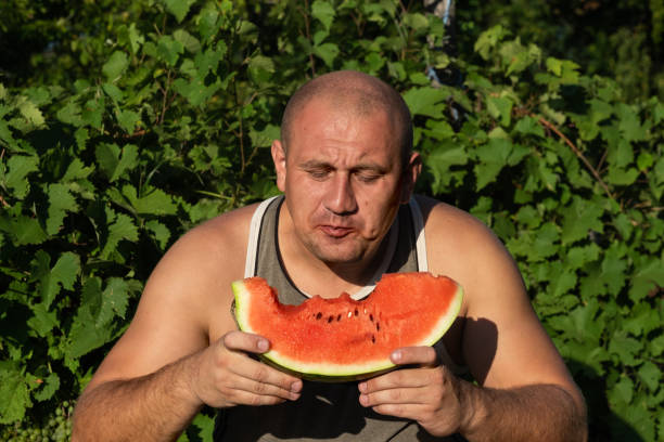 brutaler mann genießt süße rote wassermelone. 35-jährige männliche modell sitzt auf naturgrünen hintergrund und essen frisches obst. glücklich essen deich. - men 35 40 years male 30s stock-fotos und bilder