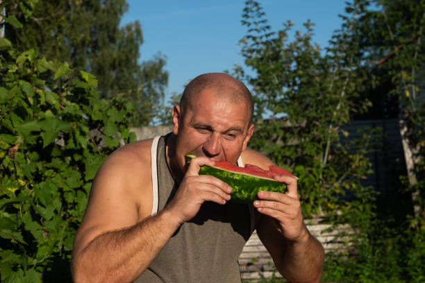 brutaler mann genießt süße rote wassermelone. 35-jährige männliche modell sitzt auf naturgrünen hintergrund und essen frisches obst. glücklich essen deich. - men 35 40 years male 30s stock-fotos und bilder