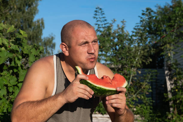 brutaler mann genießt süße rote wassermelone. 35-jährige männliche modell sitzt auf naturgrünen hintergrund und essen frisches obst. glücklich essen deich. - men 35 40 years male 30s stock-fotos und bilder