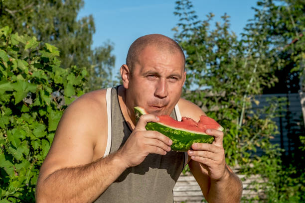 brutaler mann genießt süße rote wassermelone. 35-jährige männliche modell sitzt auf naturgrünen hintergrund und essen frisches obst. glücklich essen deich. - men 35 40 years male 30s stock-fotos und bilder