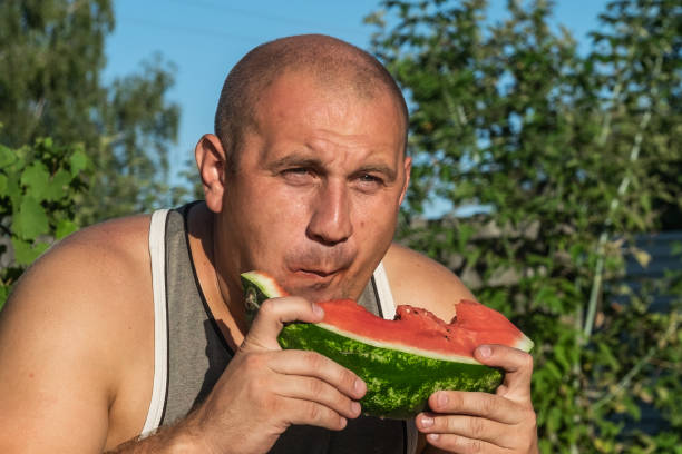 brutaler mann genießt süße rote wassermelone. 35-jährige männliche modell sitzt auf naturgrünen hintergrund und essen frisches obst. glücklich essen deich. - men 35 40 years male 30s stock-fotos und bilder