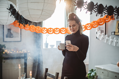 Mature woman in costume decorating home for Halloween, she is done, drinking coffee and texting with family when they will arrive