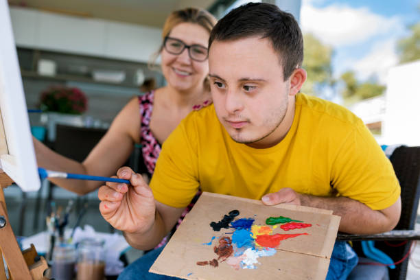 Teenager artist with Down syndrome Teen boy with Down syndrome painting on canvas with his tutor    on terrace above riverbank disabled adult stock pictures, royalty-free photos & images