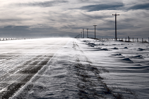 A cold and frozen winter scene of snow being blown across an asphalt highway on a cloudy overcast winter day on the Canadian Prairies.
