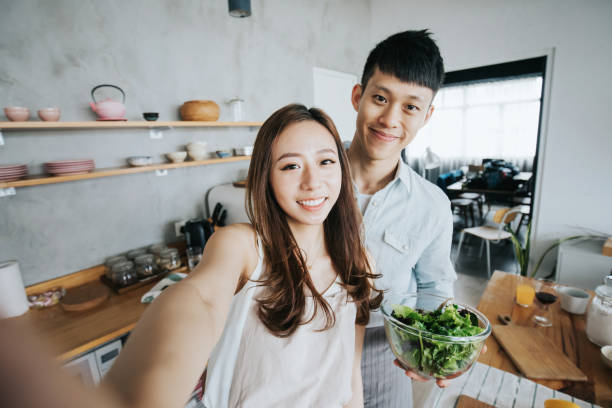 Affectionate young smiling Asian couple taking a selfie with smartphone while cooking and preparing food in kitchen together