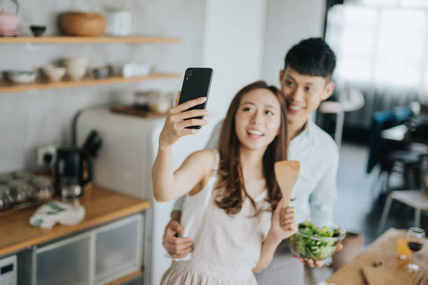 istock Affectionate young smiling Asian couple taking a selfie with smartphone while cooking and preparing food in kitchen together 1180444918