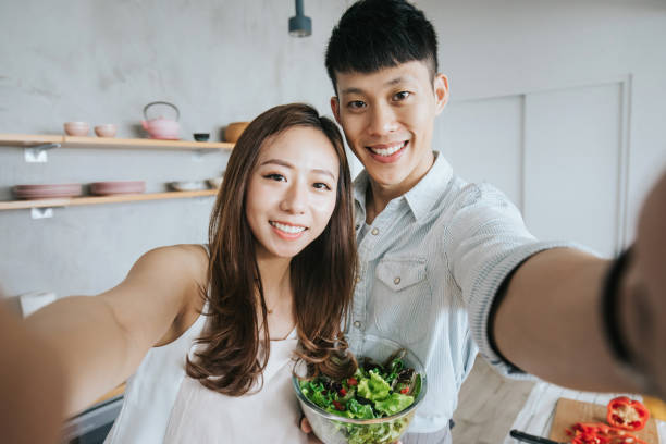 istock Affectionate young smiling Asian couple taking a selfie with smartphone while cooking and preparing food in kitchen together 1180444950