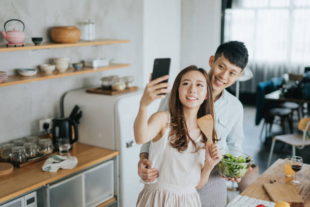 istock Affectionate young smiling Asian couple taking a selfie with smartphone while cooking and preparing food in kitchen together 1180444970