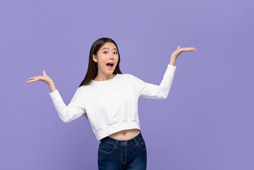 Beautiful excited young Asian woman doing presenting gesture with both hands open isolated on purple background
