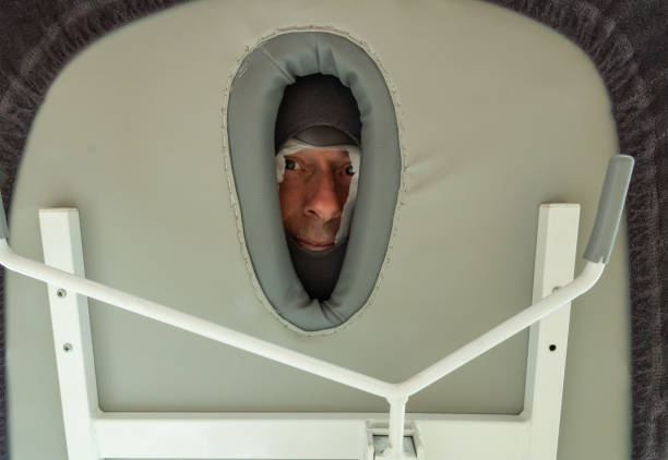 マッサージ中の理学療法士の患者 - 病院やリハビリテーションセンターのマッサージ師の頭部の穴を通してマッサージテーブルの下から見た男性の顔 - マッサージ台 ストックフォトと画像