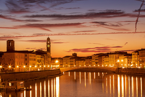 Pisa, Tuscany region, Italy. The bridge Ponte Di Mezzo over the Arno River at golden sunset in the evening.  A view of historical center. Amazing reflection of lantern lights in river Arno