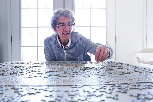A close up of an elderly woman working on a jigsaw puzzle as light from windows in the background fills the room with a bright light.