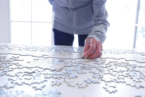 A close up of an elderly woman working on a jigsaw puzzle as light from windows in the background fills the room with a brightlight.