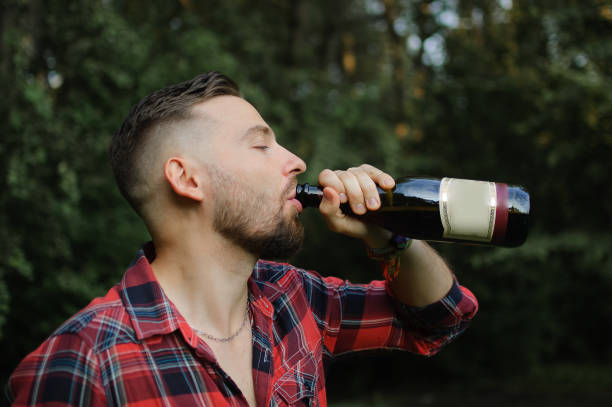 istock Brutal bearded hipster man drinking wine from the bottle standing outdoors on green trees background. Unhealthy lifestyle. 1180478098