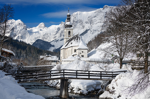 St. Sebastian church next to a mountain stream with a bridge and Reiter Alps mountains in the background, covered in snow under a blue sky, creating a winter wonderland landscape in Ramsau bei Berchtesgaden in Bavaria, Germany.